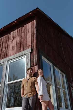 Couple posing against a red barn with large windows on a sunny day.