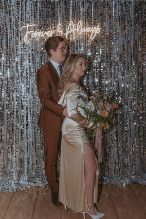 A couple embraces in front of a glittering silver backdrop with illuminated 'Forever & Always' sign.