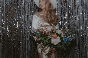 A bride in white hat and silk dress holds a colorful bouquet against a shimmering backdrop.