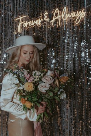 A person in a white hat holds a lush bouquet of roses against a silver sequined background.