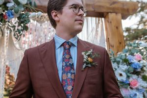 A groom in a brown suit and floral tie stands before a decorated wedding arch.