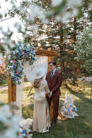 A romantic wedding ceremony setup with blue flowers and white draping against a sunset forest backdrop.