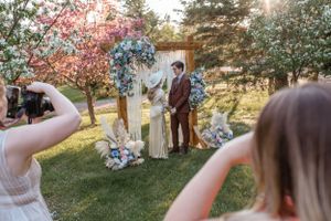 Photographers capture a wedding ceremony under a decorated wooden arch in a garden setting.