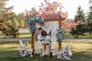 Rustic wedding ceremony setup with white macrame backdrop and blue floral arrangements in an outdoor garden setting.