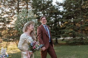 Elegant outdoor wedding scene with cream dress and brown suit against evergreen backdrop.