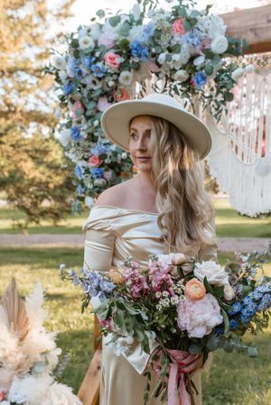 Elegant bridal portrait with cream dress, wide brim hat, and blue floral arch in garden setting.