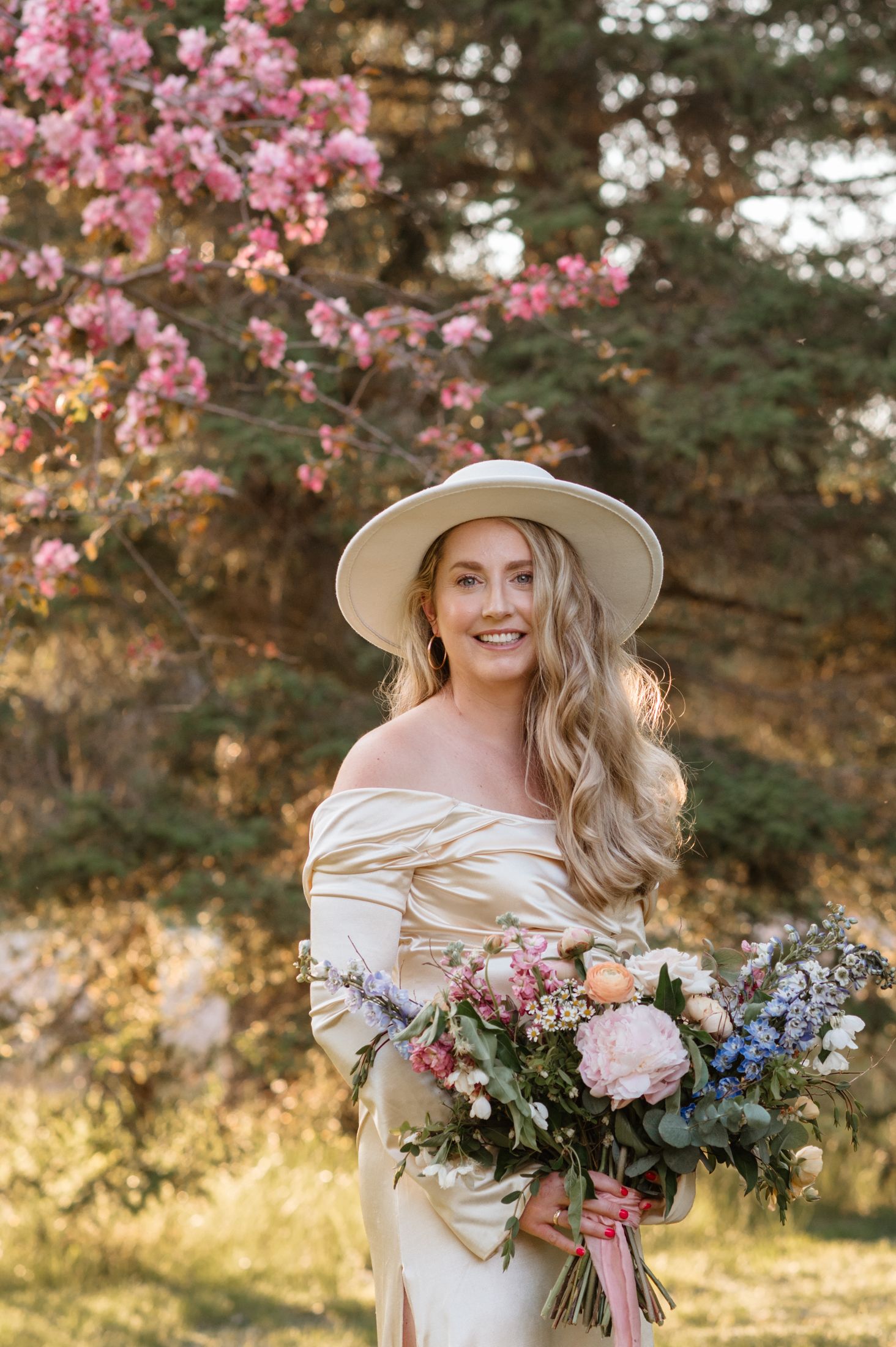 A romantic spring portrait under blooming cherry blossoms featuring an off-shoulder cream dress and wide-brimmed hat.