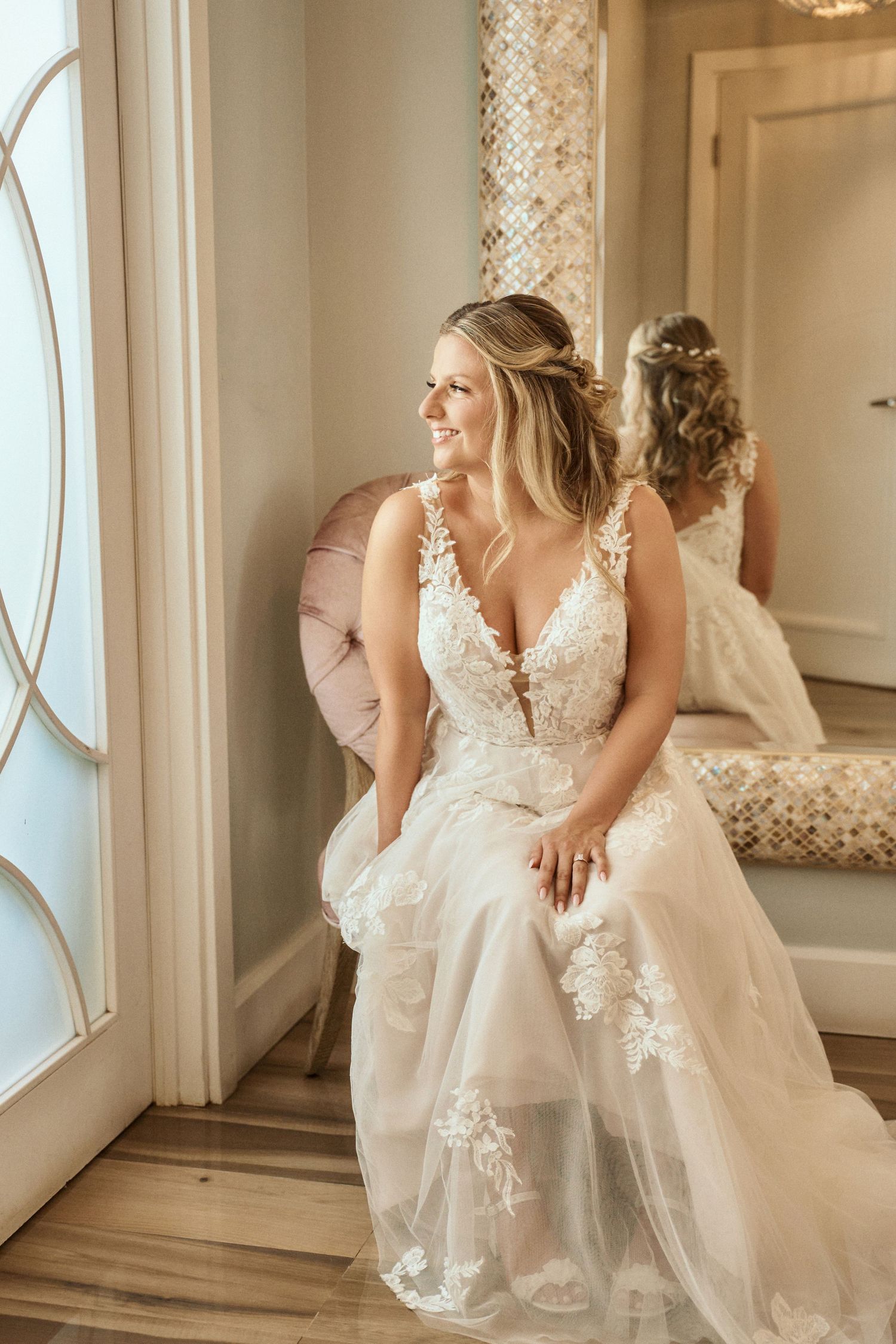 A bride in a flowing lace wedding dress poses elegantly near a window in a bright room. Jamaican Wedding Photographer