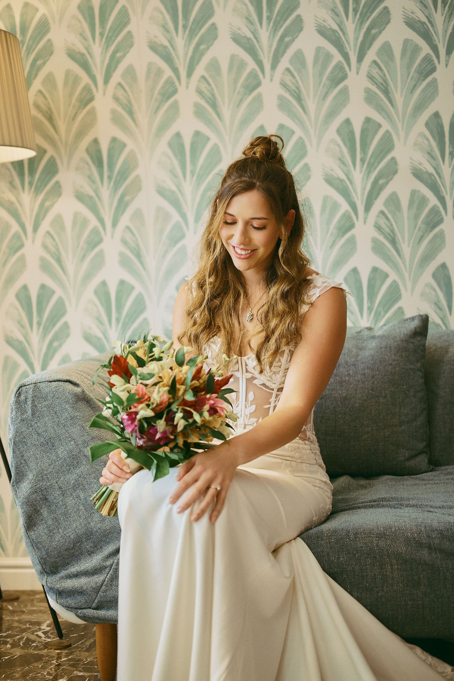 Smiling bride in white gown sits on gray sofa holding colorful bouquet, with elegant green art . Jamaican Wedding Photographer