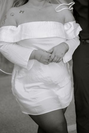 A black and white photograph of someone wearing an elegant off-shoulder white dress with ruffled details.