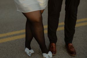 A pair of contrasting legs in white shorts and black pants meet at ground level, with one wearing white bow shoes and brown loafers.