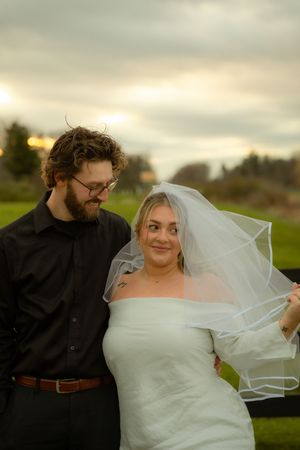 Newlywed couple poses together outdoors as bride holds her veil in the evening light.