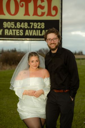 A couple poses together outdoors in formal attire, with a motel sign visible in the background.