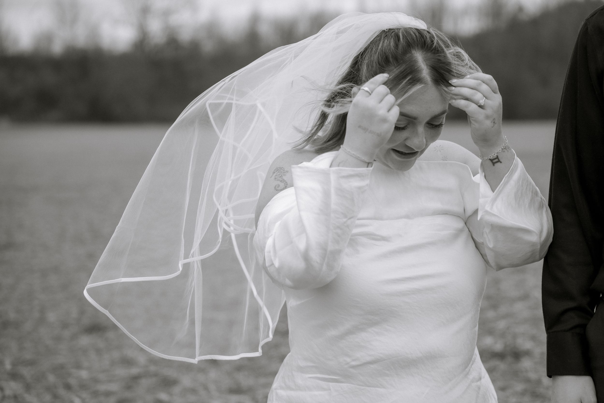 Candid black and white photo of a bride adjusting her veil outdoors in a field.