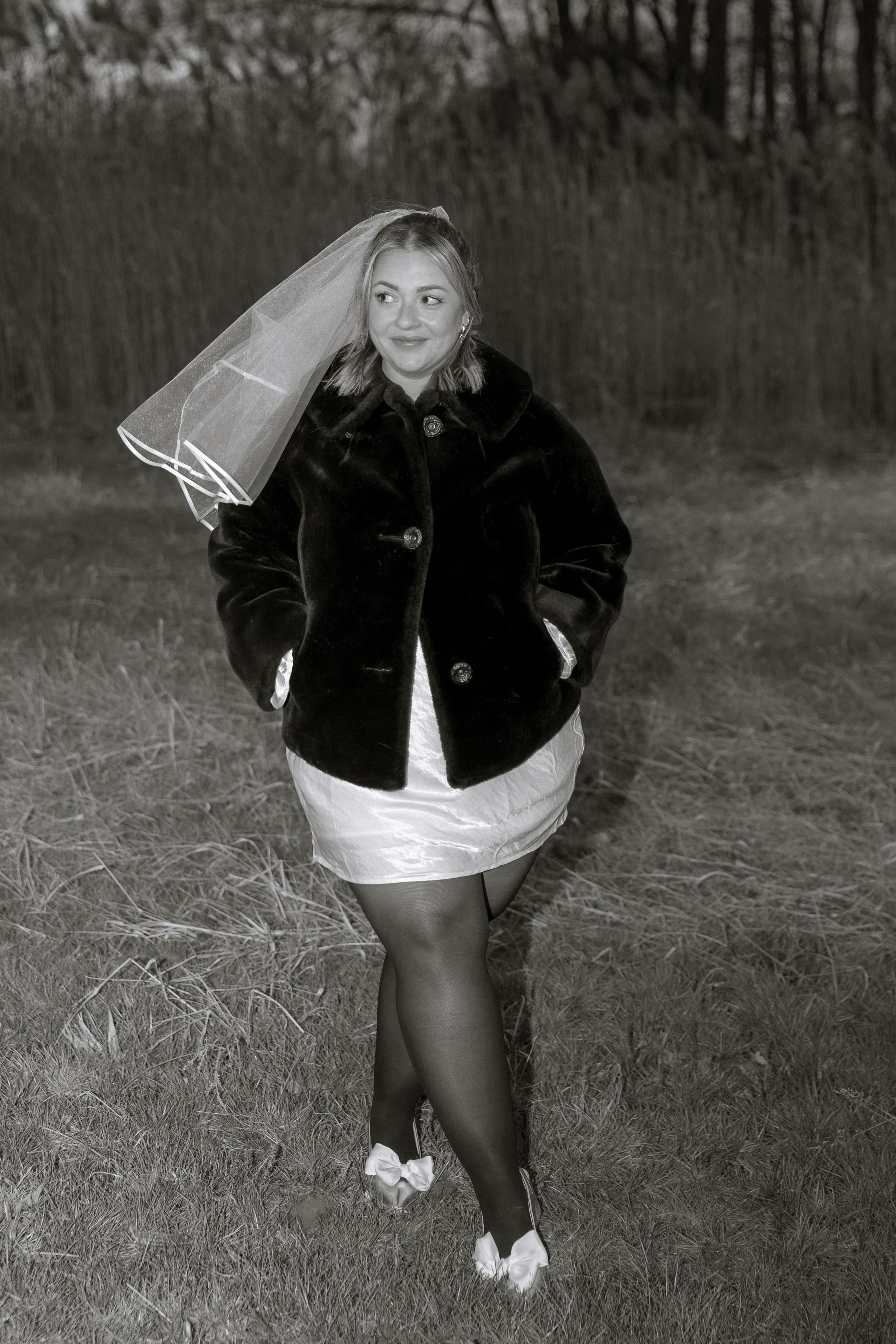 Black and white photo of someone in a dark fur coat and white dress holding up a veil in a field.
