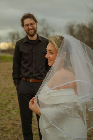 Bride in white dress with veil poses outdoors while someone in dark clothing stands behind her on a cloudy day.