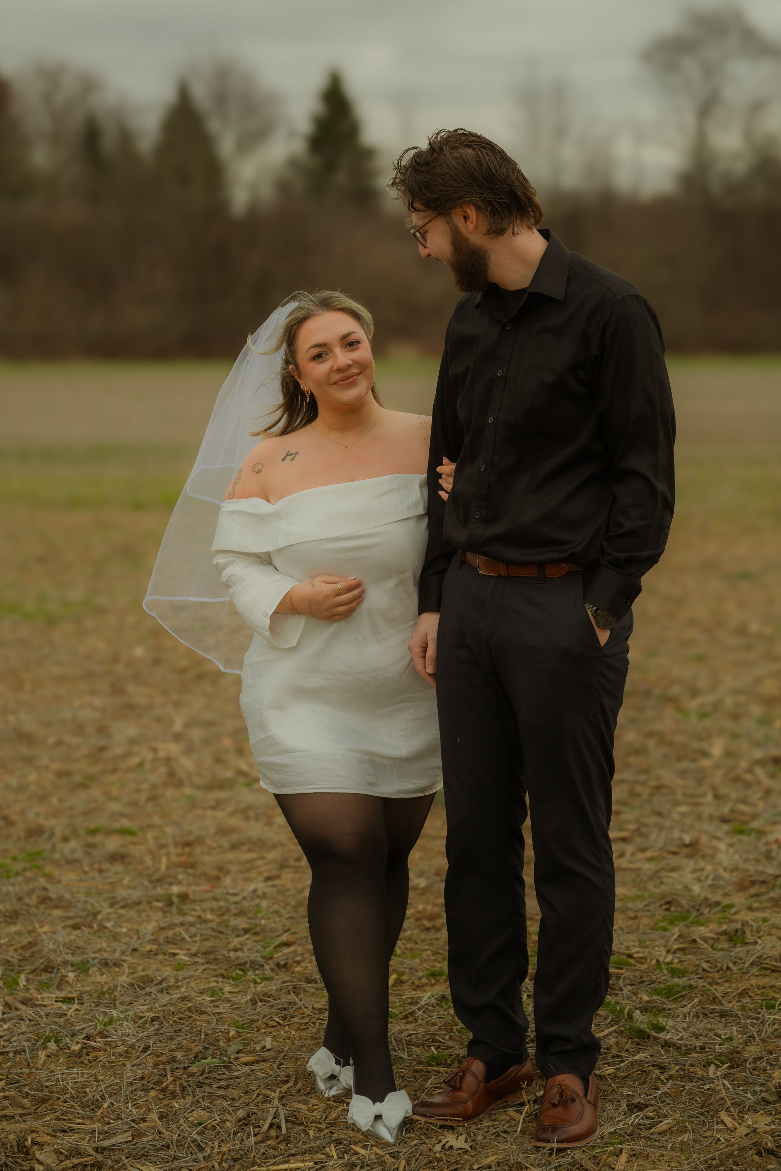 A couple poses together in a field during sunset, with the bride wearing a short white dress and veil.