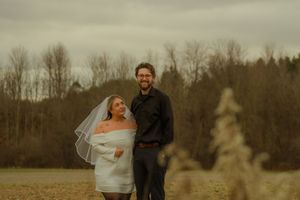 A couple stands together in a misty field at dusk with bare trees in the background.