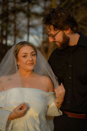 A wedding couple stands together in warm sunset light as the bride wears an off-shoulder dress with flowing veil.