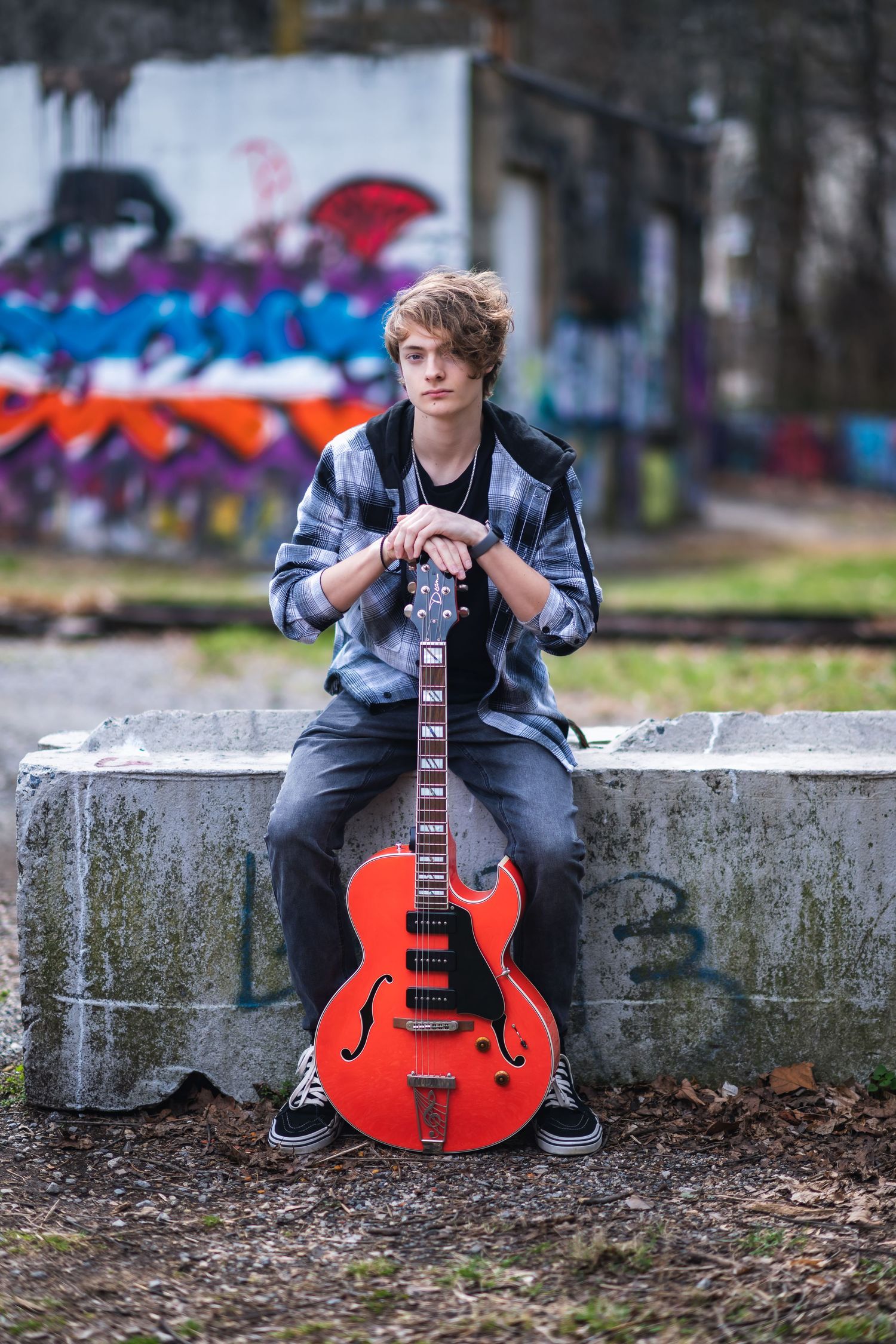 Young musician sits on concrete wall with bright red semi-hollow electric guitar against colorful graffiti backdrop.