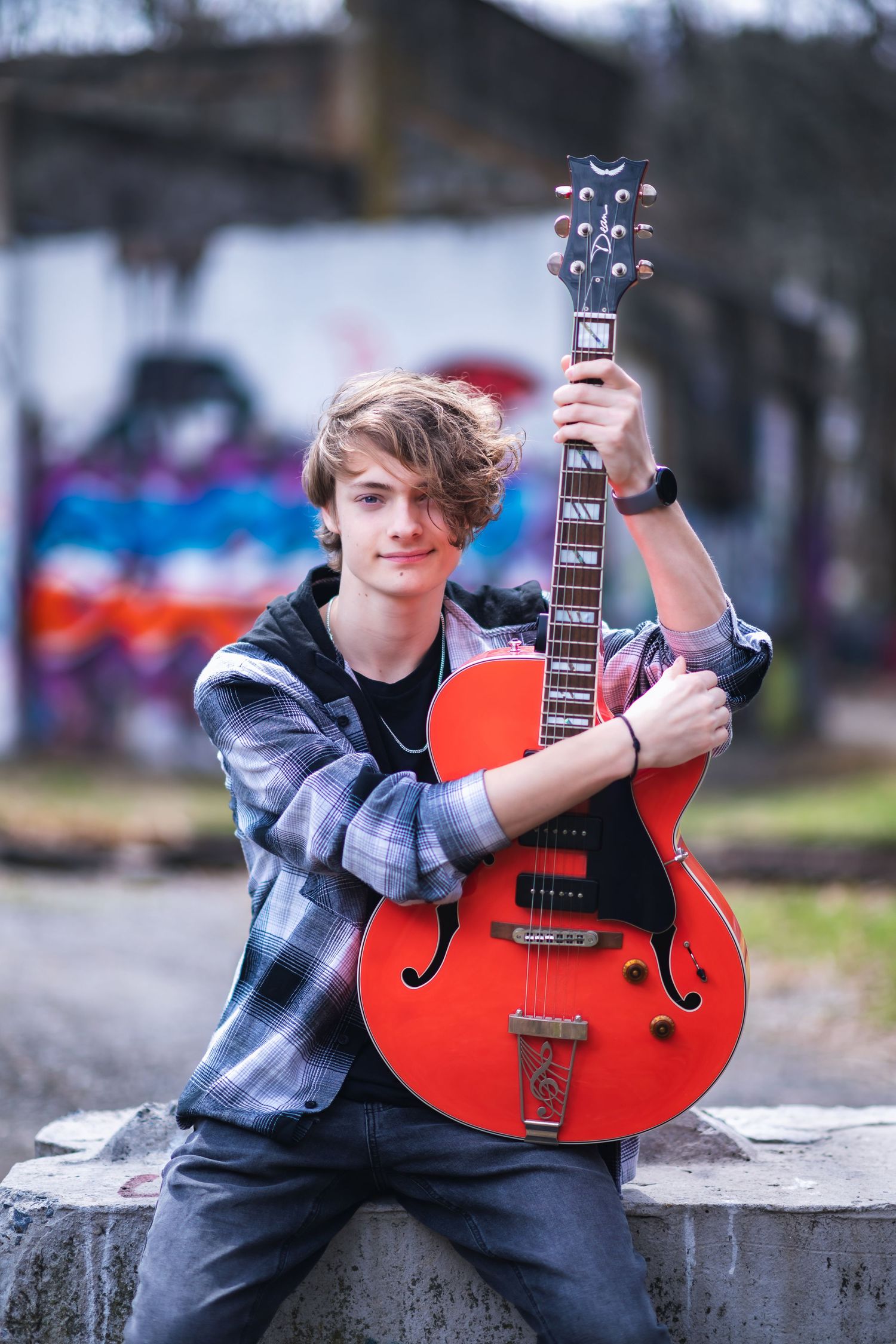 A guitarist in a plaid shirt poses outdoors with their bright red hollow-body electric guitar against a colorful background.