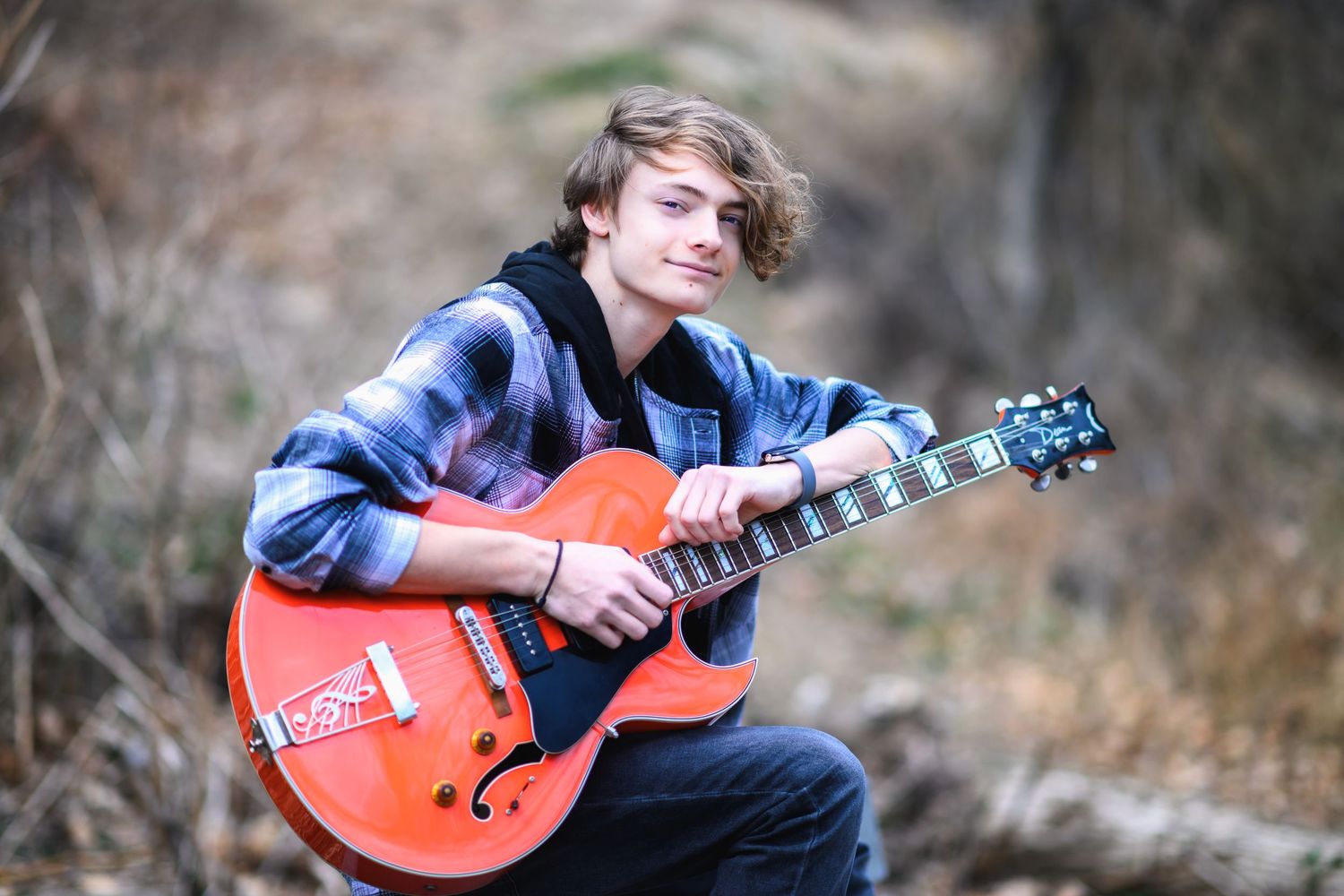 A person wearing a plaid shirt plays an orange hollow body electric guitar outdoors in nature.