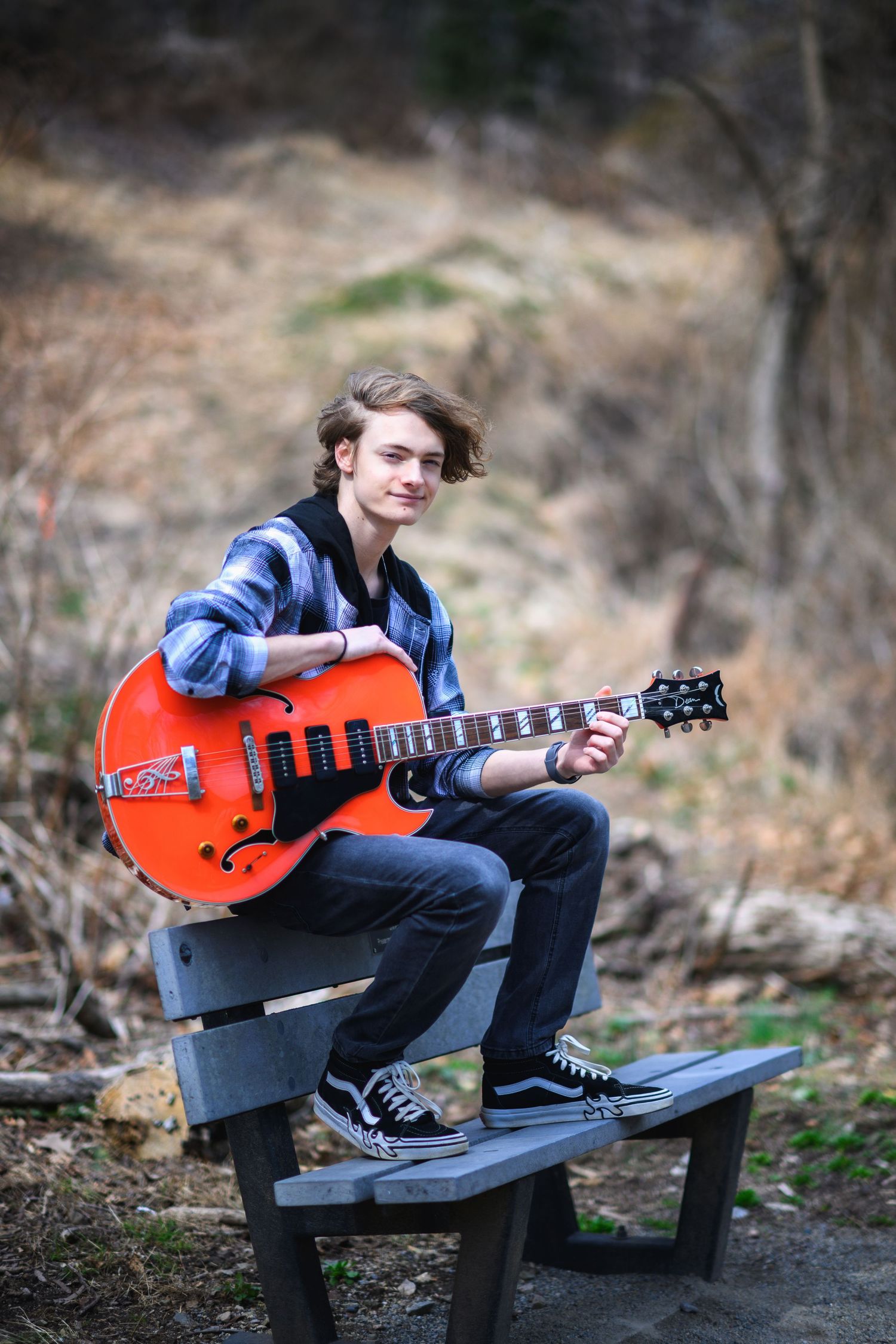 Young musician plays bright orange electric guitar while sitting on outdoor bench in casual attire.