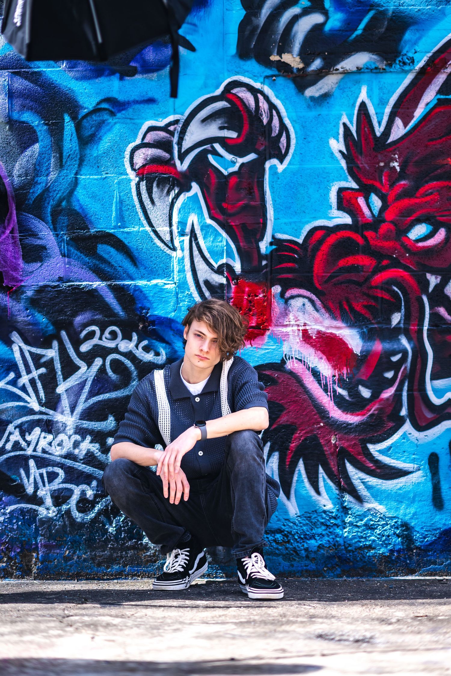 A person sits against a vibrant graffiti wall featuring red and blue dragon-like artwork in an urban setting.