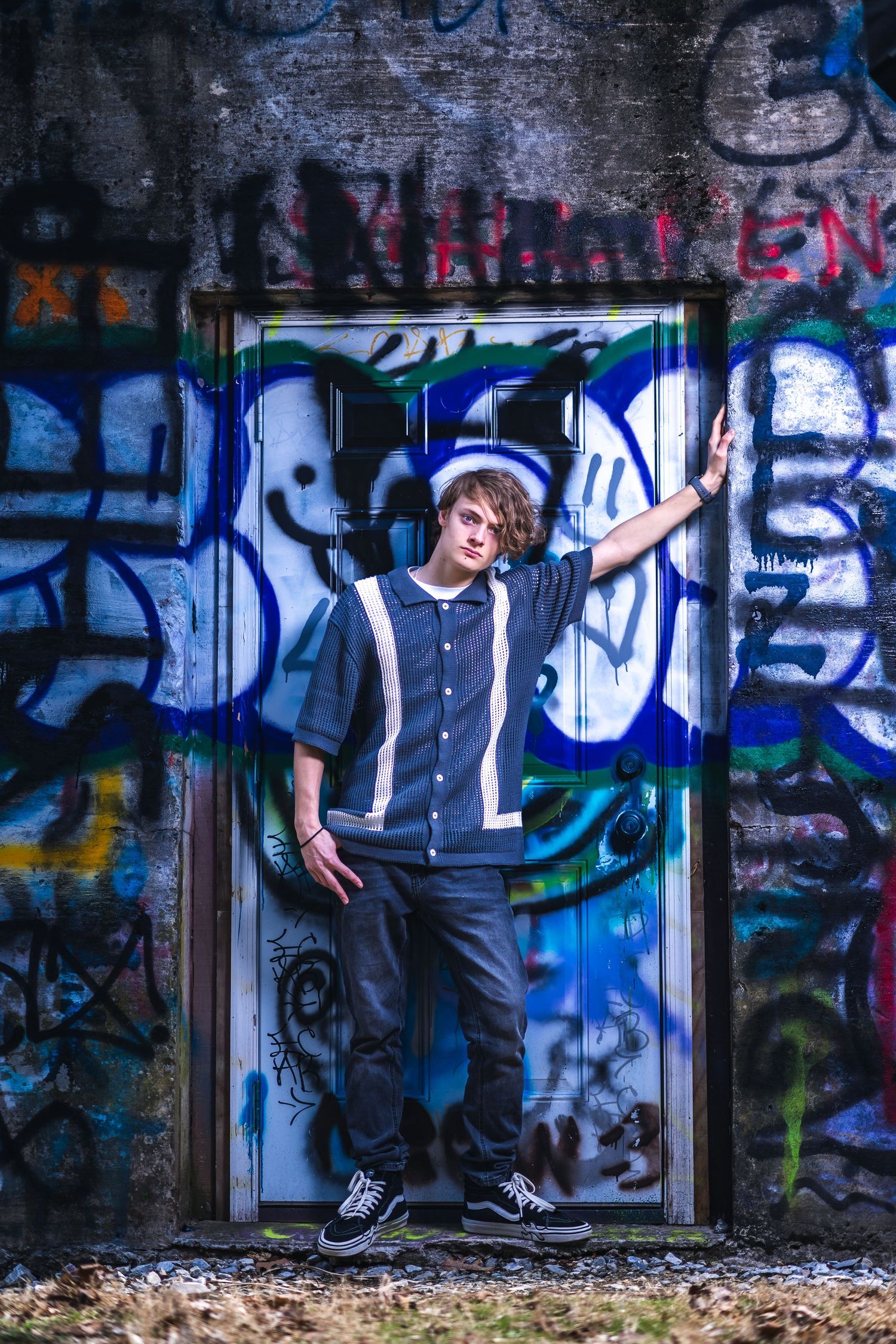 A person poses in doorway against colorful graffiti wall wearing casual streetwear and sneakers.