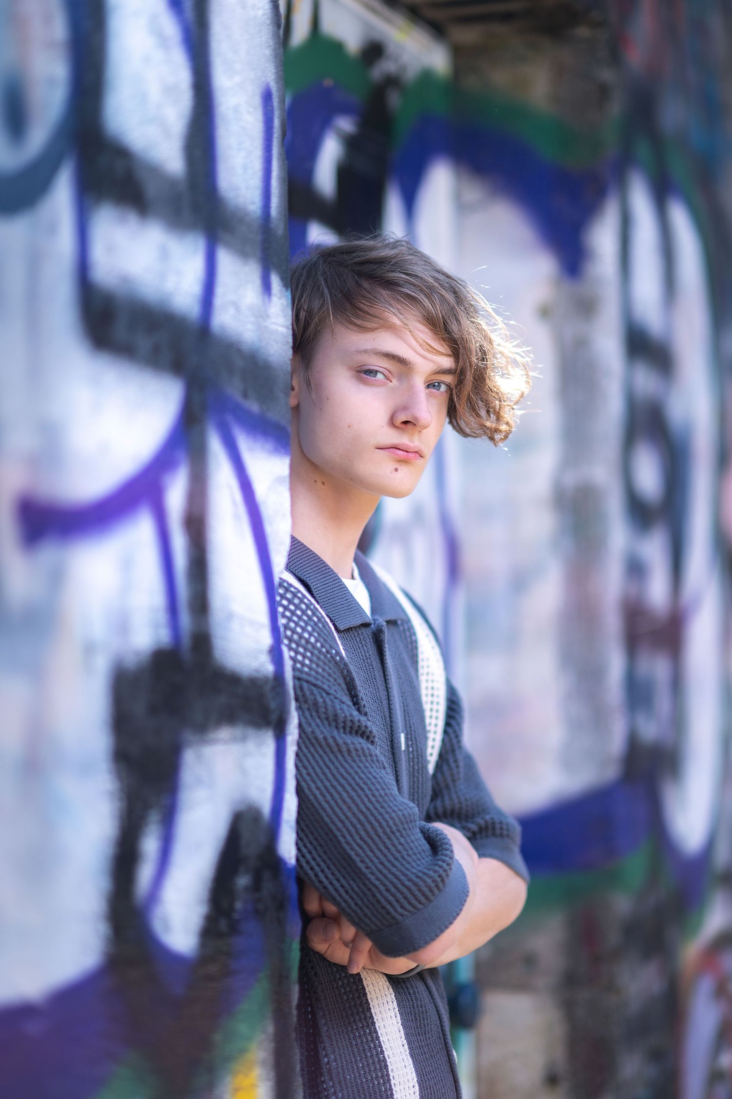 A person with windswept hair poses against a graffiti wall, wearing a dark knit sweater in an artistic portrait.