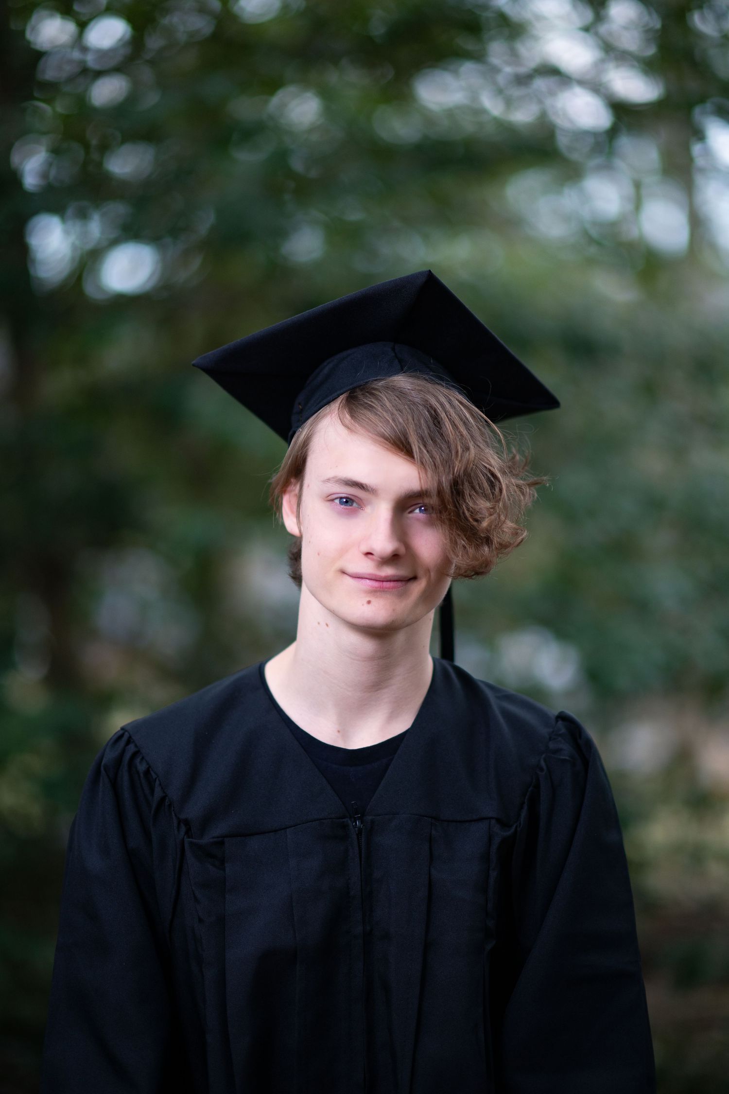 Graduate in traditional black cap and gown poses outdoors against soft blurred natural background.