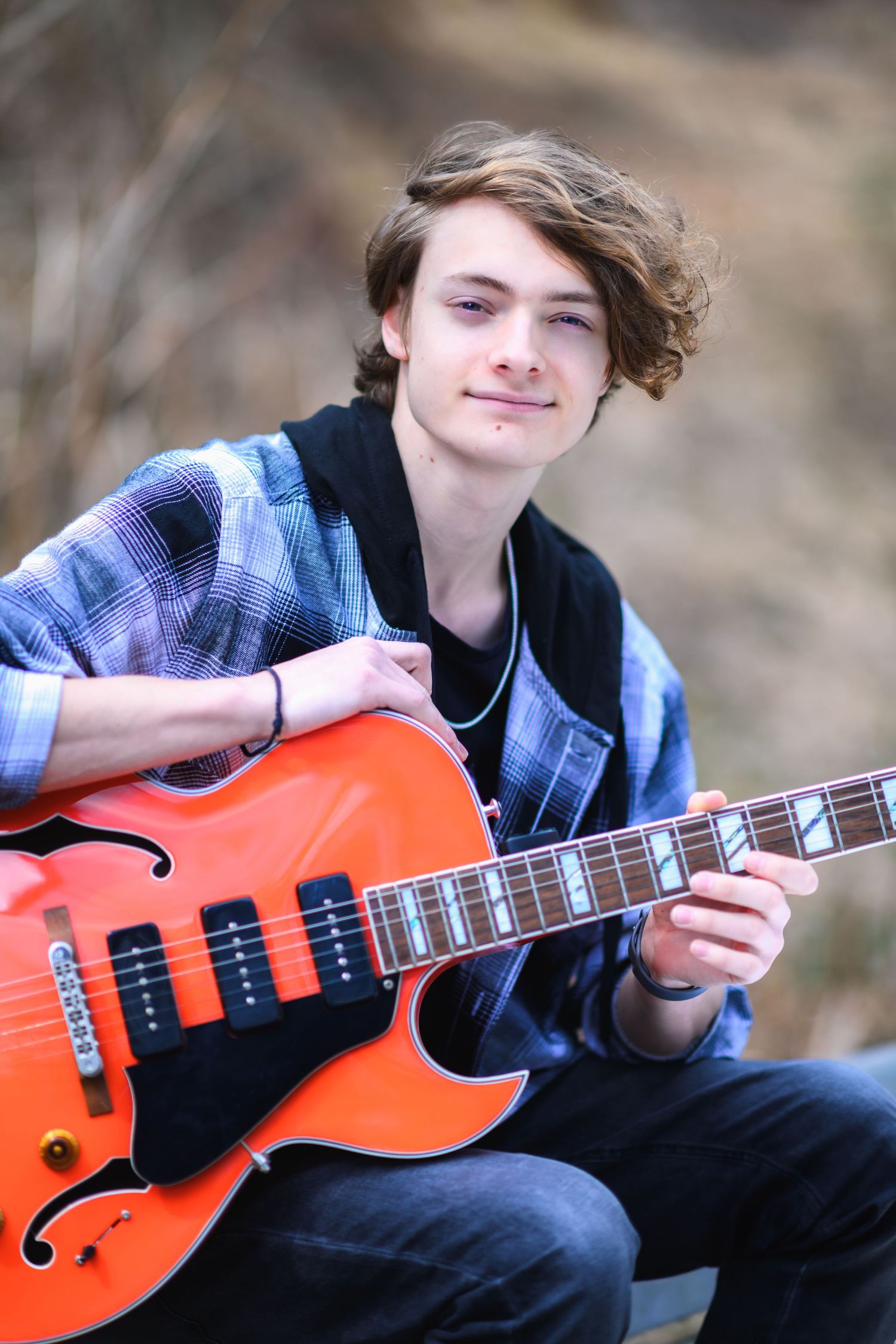 Young musician in a plaid shirt and hoodie plays an orange hollow-body electric guitar outdoors in a natural setting.
