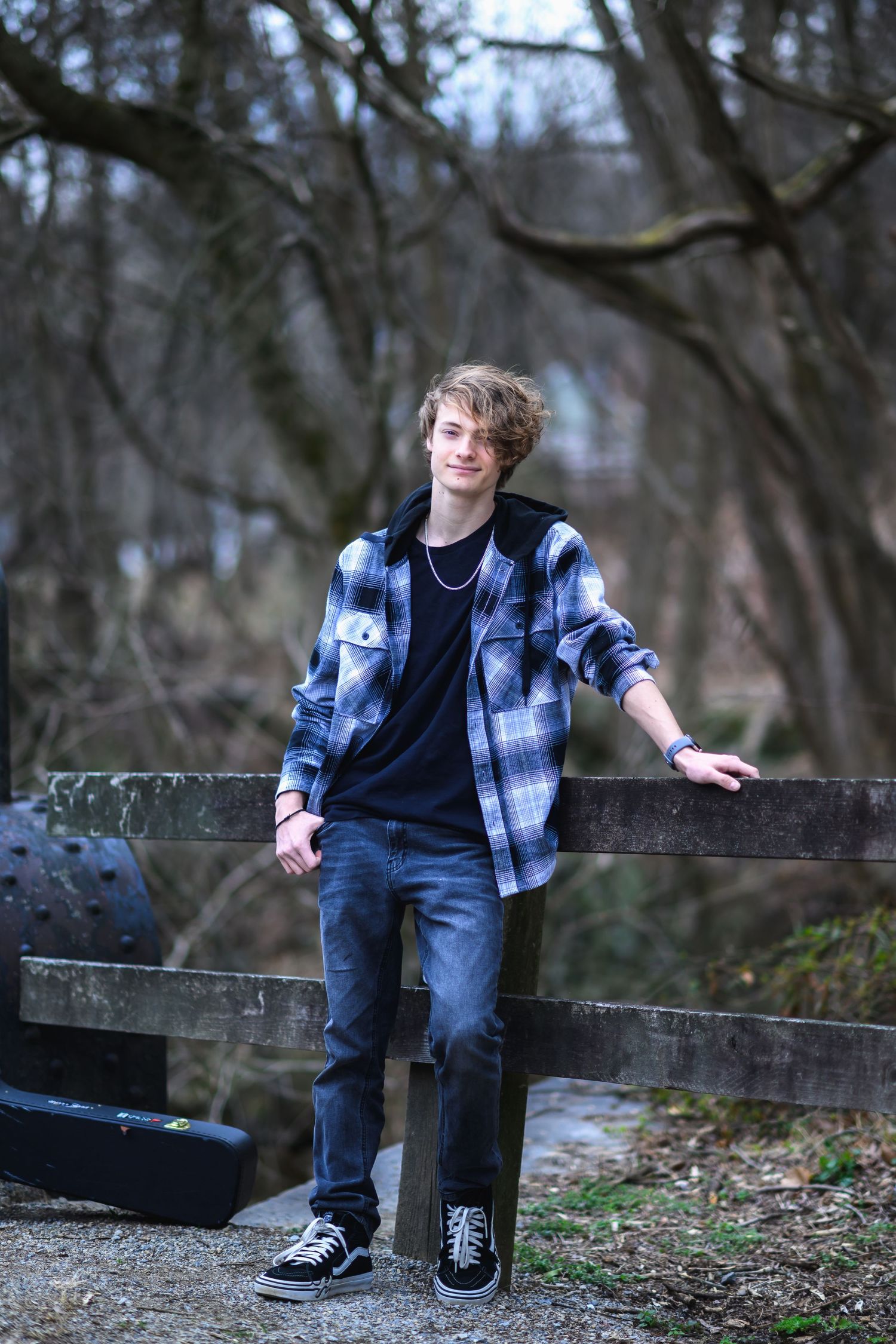 A casual outdoor portrait showing someone in a blue plaid flannel shirt and jeans leaning against a wooden fence in the woods.