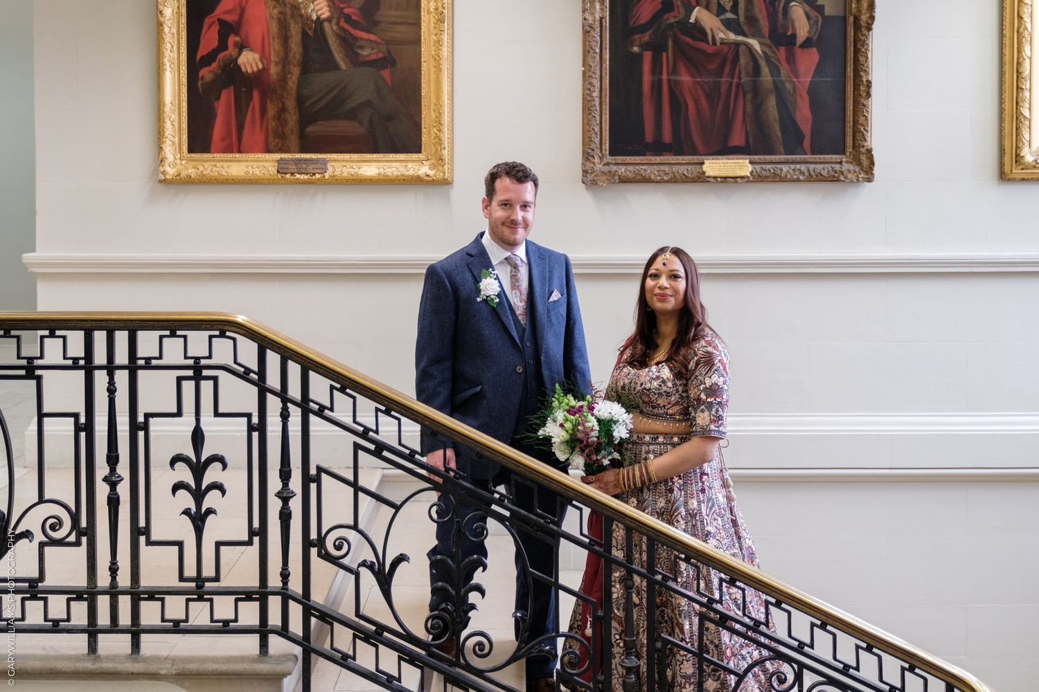A couple stands on an elegant staircase with ornate iron railings and classic paintings adorning the walls.