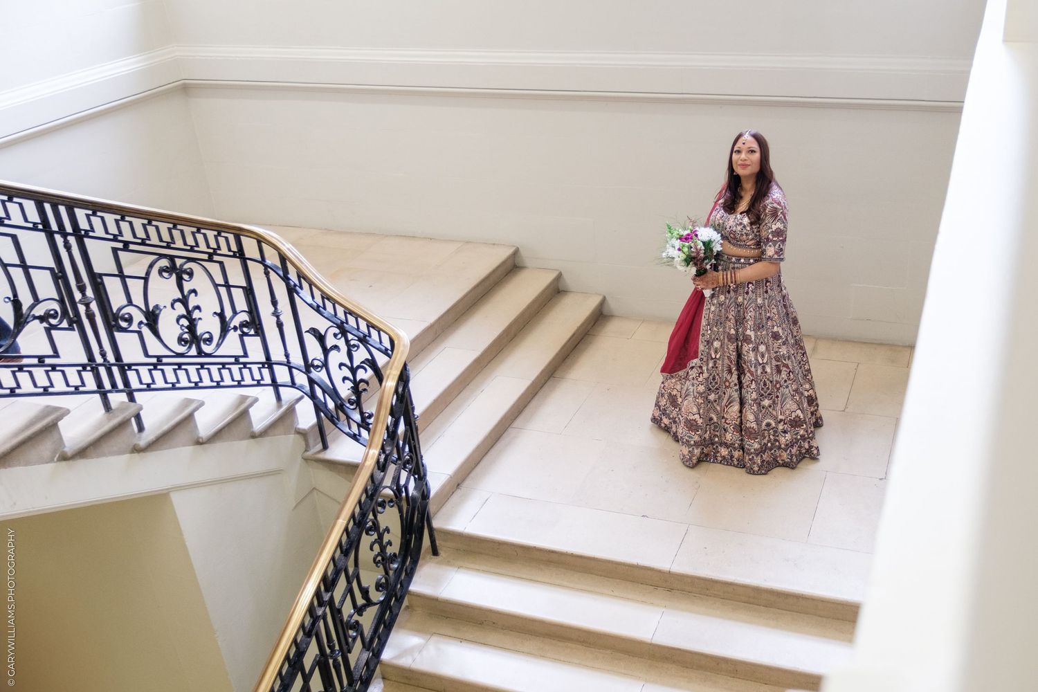 A person wearing an ornate traditional Indian lehenga stands on elegant white stairs with decorative black railings.