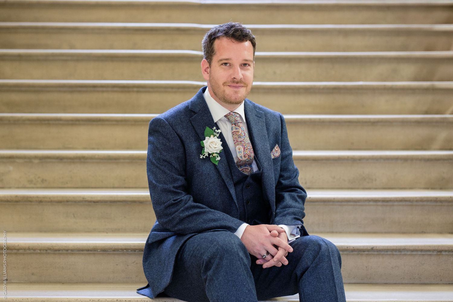 A groom in a navy suit with boutonniere sits on stone steps during a wedding photoshoot.