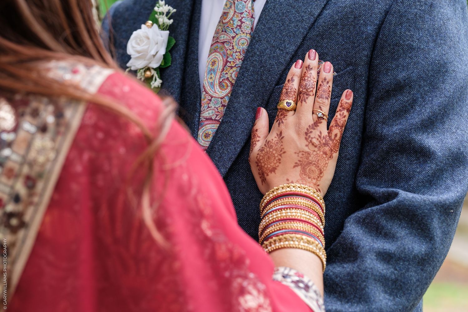 Close-up of a henna-decorated hand with bangles pressing against a navy blue blazer during a wedding ceremony.