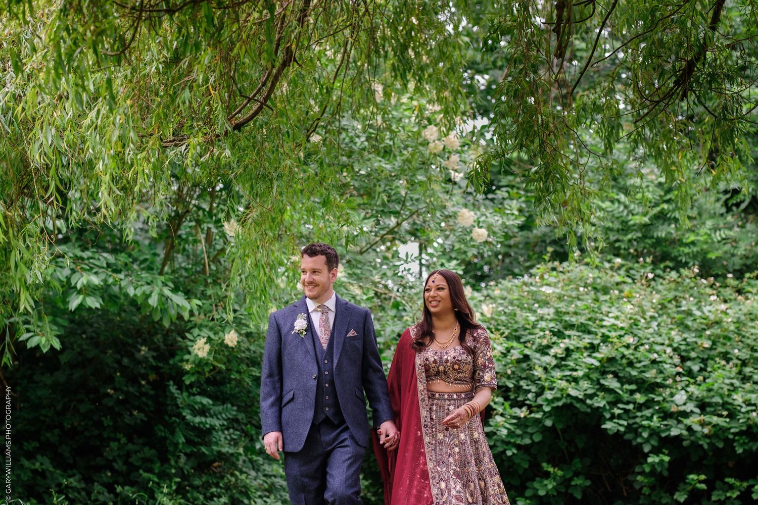 Couple walking together in garden beneath willow trees wearing formal attire.