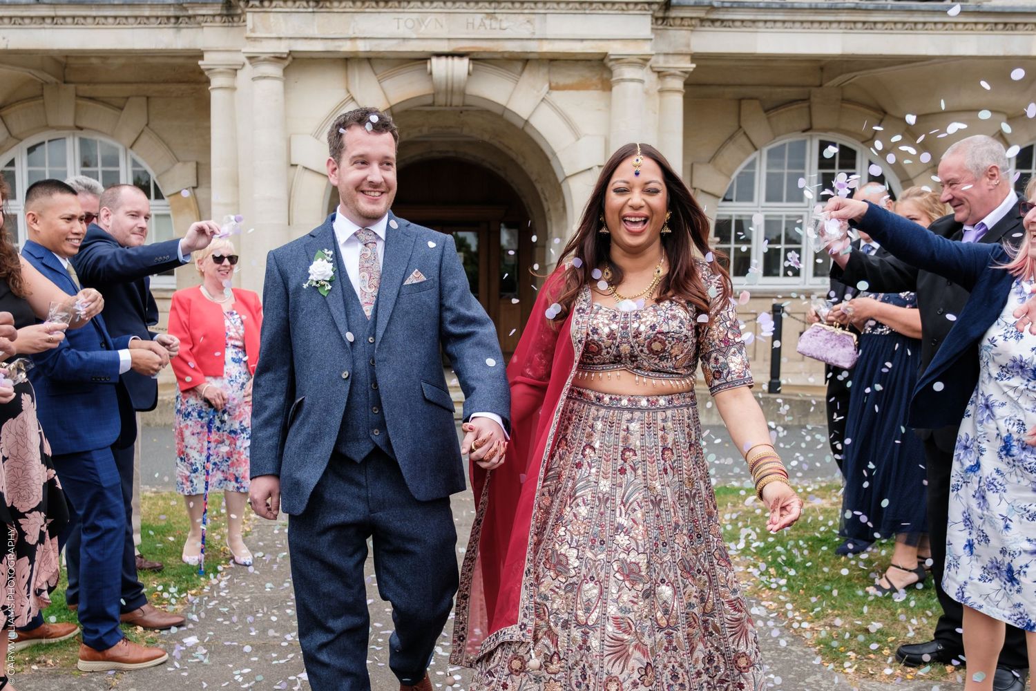Bride and groom walk through confetti while wedding guests cheer outside a grand historic building.