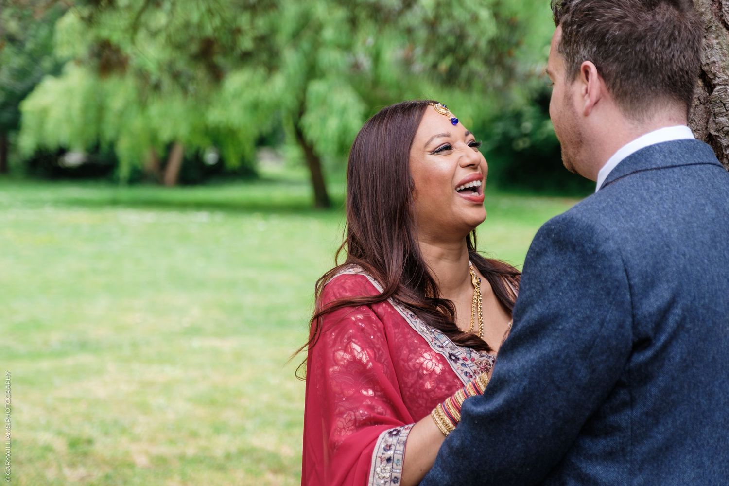 A couple laughs together on a lawn with green willow trees in the background.