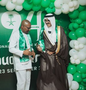 Two people interact at a Saudi National Day celebration with green and white balloon decorations and Saudi emblems in background.