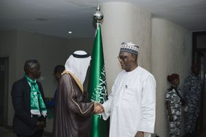 Diplomats in traditional attire shake hands during formal meeting with Saudi Arabian flag visible in background.