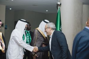 Business leaders in traditional Middle Eastern attire greet each other with handshakes at formal event with Saudi flag visible.
