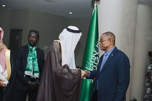 Delegates in traditional Saudi dress and business attire meet during a diplomatic event with green Saudi flags.