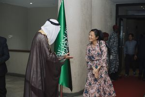 A Saudi official in traditional dress speaks with someone in a floral dress in a formal indoor setting.