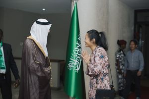 A diplomat in traditional Saudi dress meets with a woman wearing a floral print dress in front of a green Saudi flag.