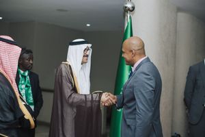 Two officials in formal attire exchange a handshake during a diplomatic meeting with a prominent Saudi Arabian flag visible.