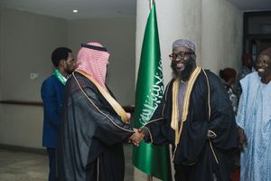 Two dignitaries in traditional Middle Eastern attire shake hands during a diplomatic meeting with Saudi flag in background.