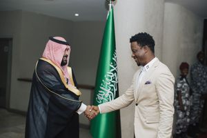 Two men in formal attire shake hands in front of a green Saudi Arabian flag during a diplomatic meeting.