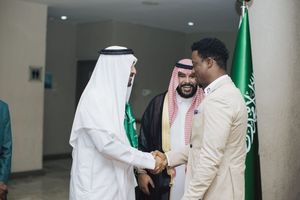 Two individuals wearing traditional Middle Eastern and Western attire shake hands in front of Saudi Arabian flag.